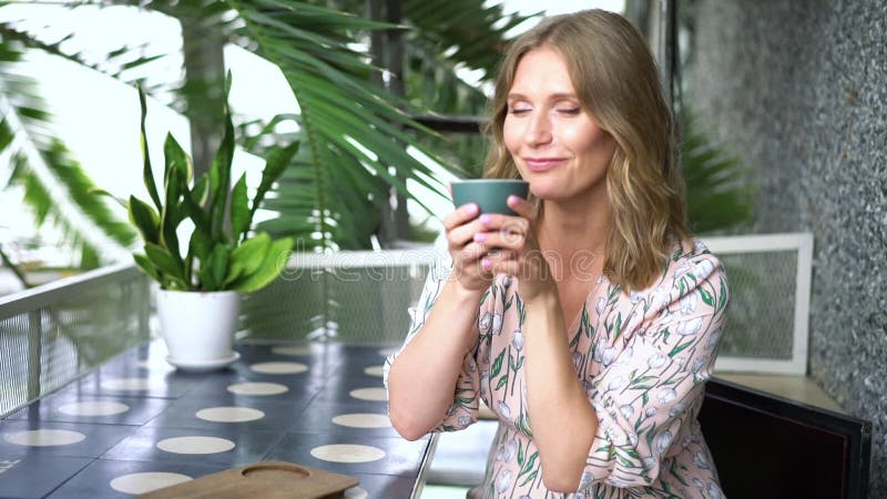 Belle femme blonde assise à une table dans un café et boit un café avec plaisir banque de vidéos