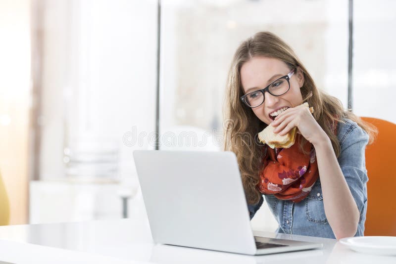 Belle femme d'affaires mangeant le sandwich tandis qu'utilisant l'ordinateur portable dans le bureau images libres de droits