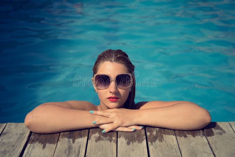 Beautiful woman relaxing at the poolside with wet hair wearing sunglasses lifted with her arms on the wood side of the pool. Beautiful woman relaxing at the poolside with wet hair wearing sunglasses lifted with her arms on the wood side of the pool