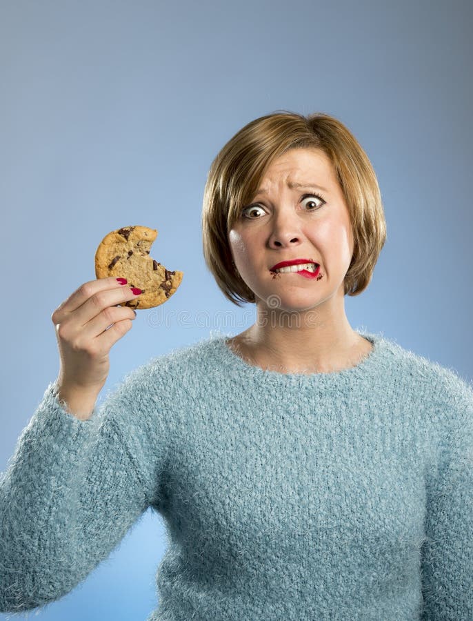 Young blond cute beautiful caucasian woman with chocolate stain in her mouth eating big delicious cookie looking with temptation thinking and if ignoring diet vs sweet junk food isolated background. Young blond cute beautiful caucasian woman with chocolate stain in her mouth eating big delicious cookie looking with temptation thinking and if ignoring diet vs sweet junk food isolated background