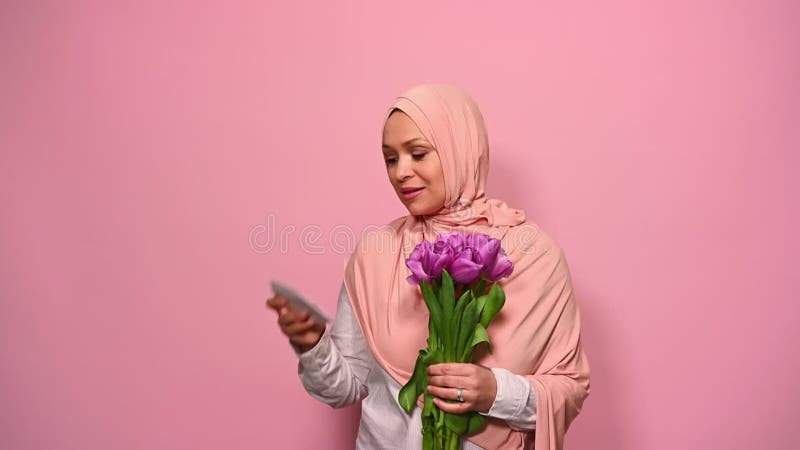 Belle femme musulmane en hijab rose lit la carte de voeux dans l'enveloppe tout en obtenant mignon bouquet de tulipes pour les fem banque de vidéos