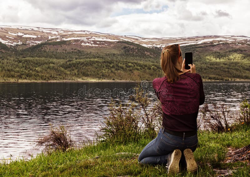 Belle femme seule s'asseyant sur le rivage d'un lac de montagne et prendre une photo sur un smartphone photos stock Belle femme seule s'asseyant sur le rivage d'un lac de montagne et prendre une photo sur un smartphone photos stock