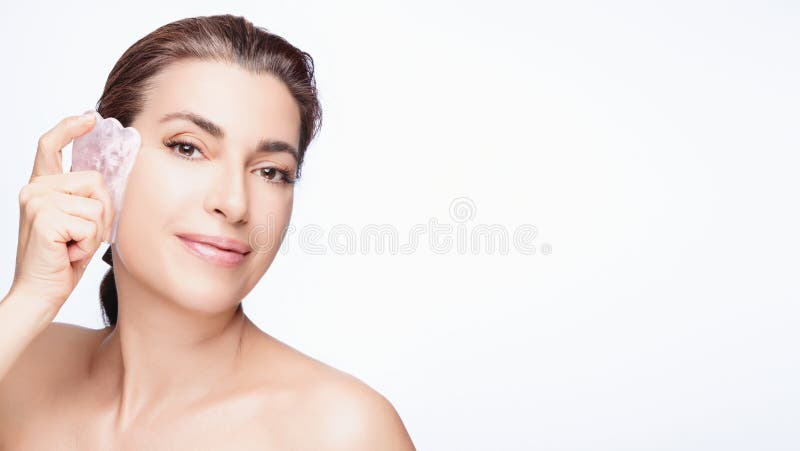 An attractive young woman with bare shoulders uses a rose quartz gua sha on her skin, looking serene and happy against a clean white background. An attractive young woman with bare shoulders uses a rose quartz gua sha on her skin, looking serene and happy against a clean white background.