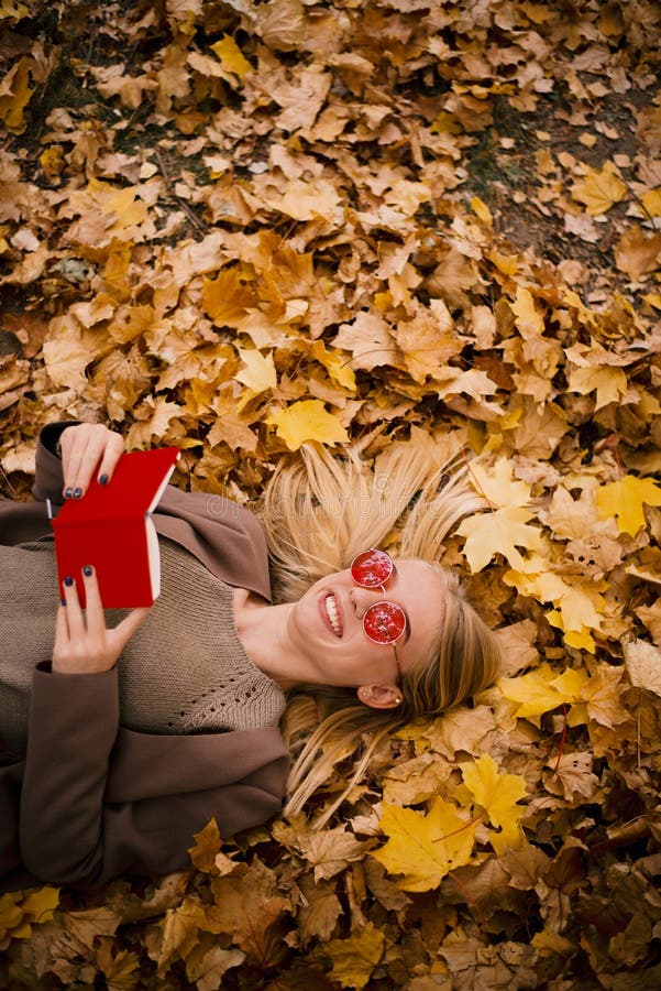 Belle jeune blonde dans des mensonges roses en verre dans des feuilles d'automne jaunes, lisant un livre dans la couverture rouge image libre de droits Belle jeune blonde dans des mensonges roses en verre dans des feuilles d'automne jaunes, lisant un livre dans la couverture rouge image libre de droits