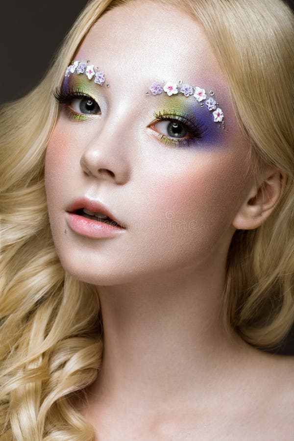 Belle jeune femme blonde avec la couleur créative, les boucles et les fleurs de maquillage sur des sourcils Visage de beauté Art photo stock Belle jeune femme blonde avec la couleur créative, les boucles et les fleurs de maquillage sur des sourcils Visage de beauté Art photo stock