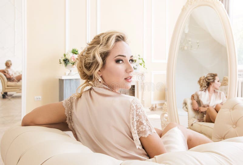 Belle jeune femme blonde sexy dans une robe longue en soie se reposant sur un sofa devant le miroir images stock Belle jeune femme blonde sexy dans une robe longue en soie se reposant sur un sofa devant le miroir images stock