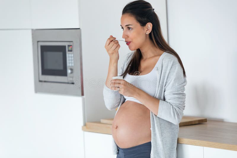 Belle jeune femme enceinte mangeant du yaourt tout en se tenant dans la cuisine à la maison photos libres de droits Belle jeune femme enceinte mangeant du yaourt tout en se tenant dans la cuisine à la maison photos libres de droits