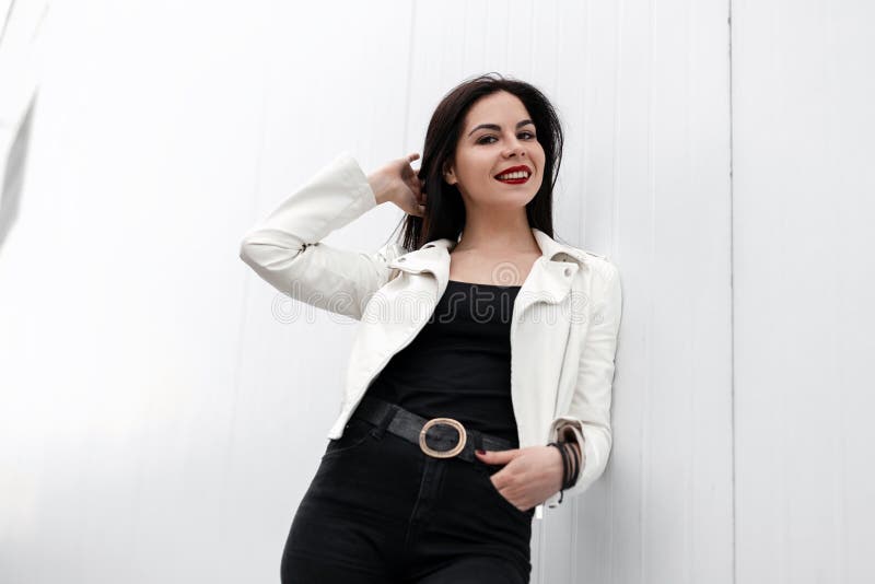 Belle jeune femme gaie avec les lèvres rouges dans une veste en cuir élégante dans un T-shirt dans des jeans noirs à la mode posa photographie stock Belle jeune femme gaie avec les lèvres rouges dans une veste en cuir élégante dans un T-shirt dans des jeans noirs à la mode posa photographie stock