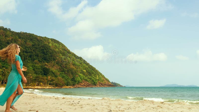 Belle jeune femme pieds nus marchant sur la plage ensoleillée robe de soie bleue flottant au vent une belle journée pour une prome clips vidéos