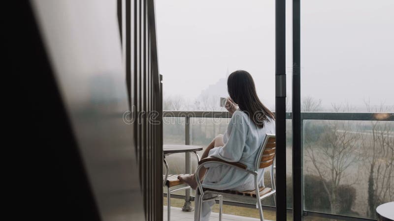 Belle jeune femme rétroviseur dans la robe de chambre ayant un café sur la terrasse de l'hôtel avec le mouvement lent de vue brume banque de vidéos
