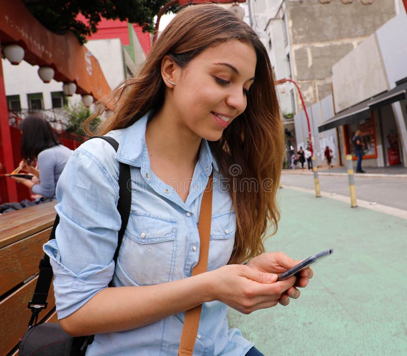 Belle jeune femme s'asseyant sur la transmission de messages de banc de rue avec le téléphone portable dans le sao Paulo City, Br image stock Belle jeune femme s'asseyant sur la transmission de messages de banc de rue avec le téléphone portable dans le sao Paulo City, Br image stock