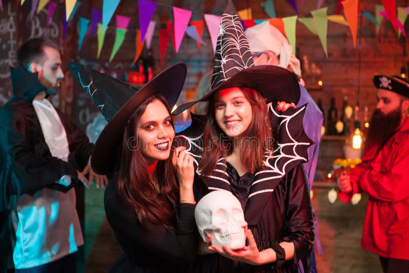 Beautiful young witch holding a human skull and smiling to the camera at halloween party. Witches looking into the camera. Beautiful young witch holding a human skull and smiling to the camera at halloween party. Witches looking into the camera.