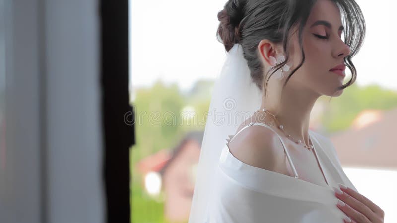 Belle mariée en robe blanche posant près d'une fenêtre lumineuse avec une expression sereine Jeune mariée portant une robe blanche banque de vidéos