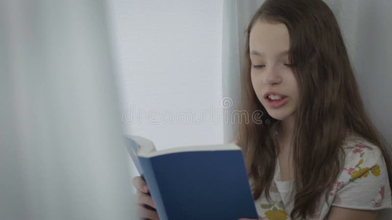 Belle petite fille lisant un livre intéressant par la fenêtre