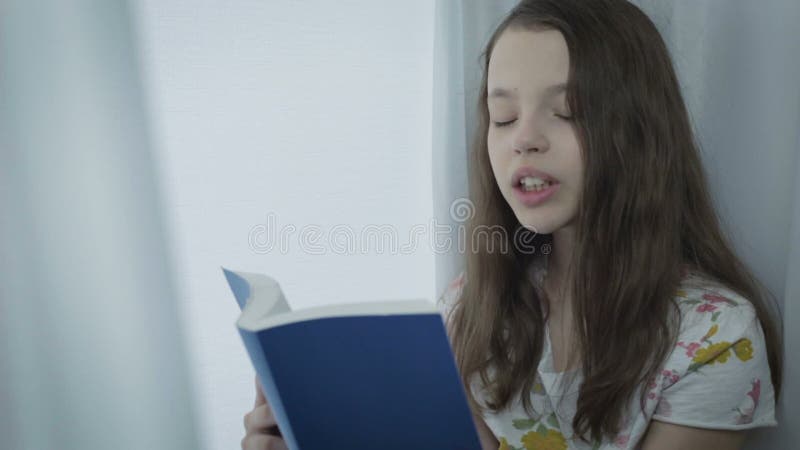 Belle petite fille lisant un livre intéressant par la fenêtre