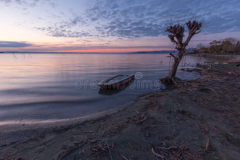 Belle vue de lac Ombrie Trasimeno au crépuscule, avec un petit bateau près d'un arbre, parfaitement toujours d'une eau et d'un a photographie stock Belle vue de lac Ombrie Trasimeno au crépuscule, avec un petit bateau près d'un arbre, parfaitement toujours d'une eau et d'un a photographie stock