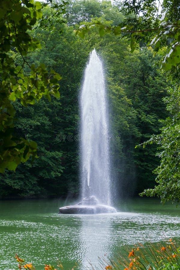 Belle vue par derrière le feuillage vert des arbres à la fontaine au milieu de l'étang avec un courant clair photographie stock libre de droits Belle vue par derrière le feuillage vert des arbres à la fontaine au milieu de l'étang avec un courant clair photographie stock libre de droits