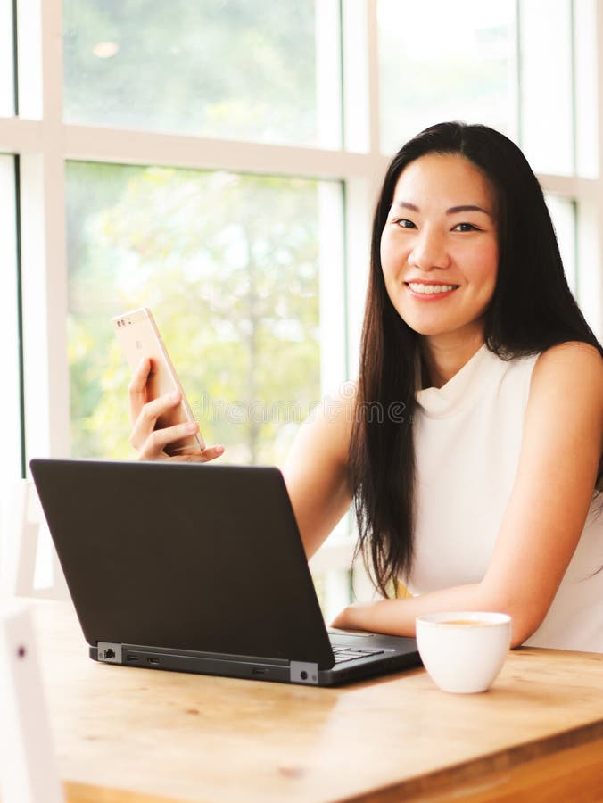 Bellissima donna asiatica seduta col computer portatile che tiene il cellulare e sorride alla macchina fotografica fotografie stock Bellissima donna asiatica seduta col computer portatile che tiene il cellulare e sorride alla macchina fotografica fotografie stock