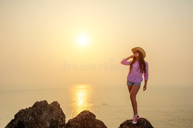 Bello castana con le gambe lunghe in cappello di paglia e breve denim mette sta sulla grande pietra contro il tramonto sopra l'oc fotografie stock libere da diritti Bello castana con le gambe lunghe in cappello di paglia e breve denim mette sta sulla grande pietra contro il tramonto sopra l'oc fotografie stock libere da diritti