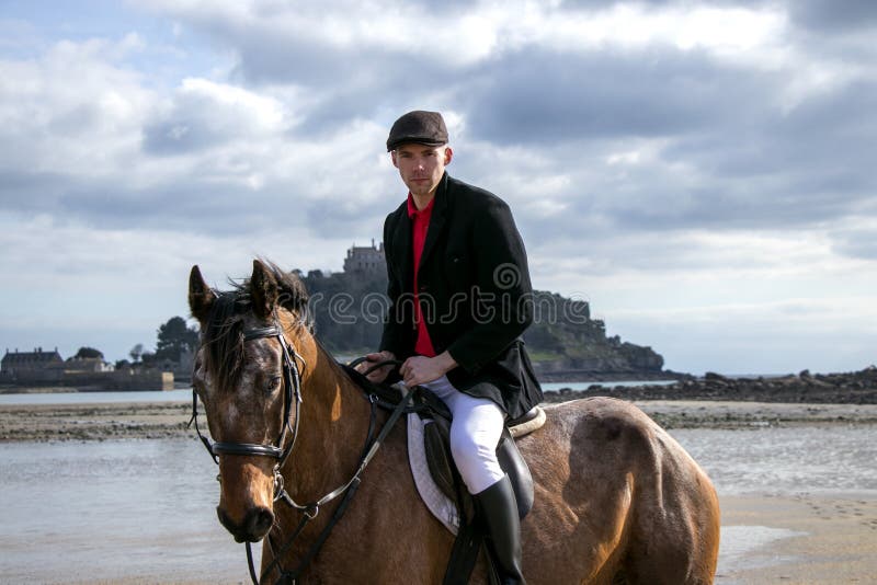 Bello cavallo da equitazione del cavaliere del cavallo maschio sulla spiaggia in abbigliamento tradizionale di guida con il suppo immagine stock libera da diritti Bello cavallo da equitazione del cavaliere del cavallo maschio sulla spiaggia in abbigliamento tradizionale di guida con il suppo immagine stock libera da diritti