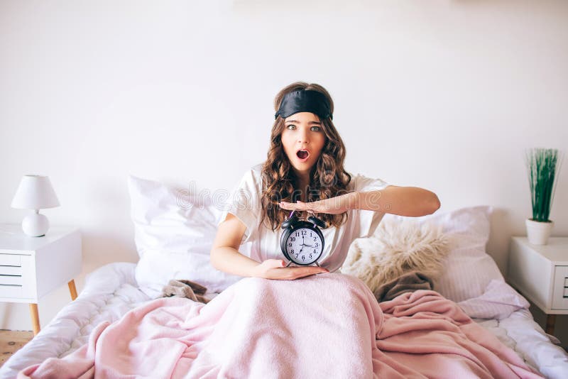 Bello giovane castana moro sveglia nel suo letto Orologio confuso della tenuta della donna in mani Ha svegliato più successivamen fotografia stock Bello giovane castana moro sveglia nel suo letto Orologio confuso della tenuta della donna in mani Ha svegliato più successivamen fotografia stock