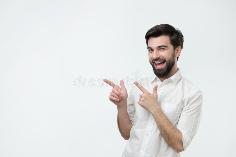 Bello tipo barbuto felice che esamina macchina fotografica, sorridente ed indicante da parte con la mano immagini stock libere da diritti Bello tipo barbuto felice che esamina macchina fotografica, sorridente ed indicante da parte con la mano immagini stock libere da diritti