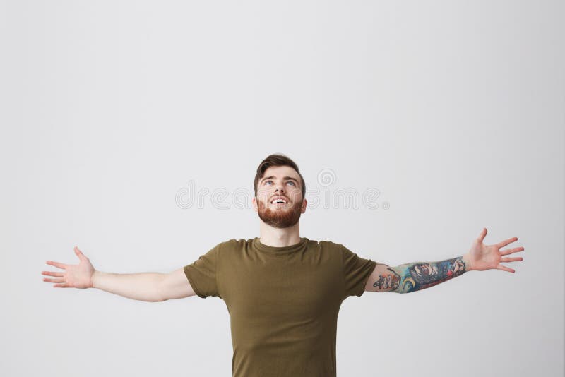 Bello uomo europeo barbuto con il braccio ed i capelli scuri tatooed in mani di diffusione della maglietta verde oliva casuale am immagine stock libera da diritti Bello uomo europeo barbuto con il braccio ed i capelli scuri tatooed in mani di diffusione della maglietta verde oliva casuale am immagine stock libera da diritti