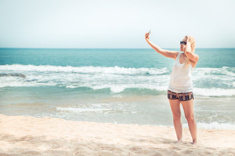 Bello viaggiatore solo della donna sulla spiaggia che prende il selfie dell'immagine sul telefono durante la vacanza di feste di fotografia stock libera da diritti Bello viaggiatore solo della donna sulla spiaggia che prende il selfie dell'immagine sul telefono durante la vacanza di feste di fotografia stock libera da diritti