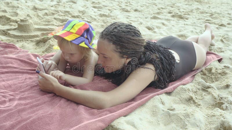 Bemuttern Sie Spiele mit der kleinen Tochter, die Smartphone auf dem Strand verwendet