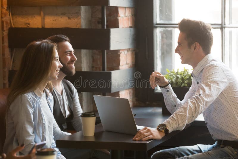 Male architect talking to smiling millennial couple, consulting on housing project, realtor explaining mortgage and contract condition to excited young couple during friendly meeting in coffee shop. Male architect talking to smiling millennial couple, consulting on housing project, realtor explaining mortgage and contract condition to excited young couple during friendly meeting in coffee shop