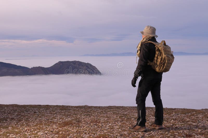 BERGABENADVENTURER MIT RÜCKPACKUNG AUF DEM GIPFEL EINES BERGES, DER EIN Tal BEOBACHTET, DAS VON EINEM DENSE FOG GEDECKT WIRD stockbild