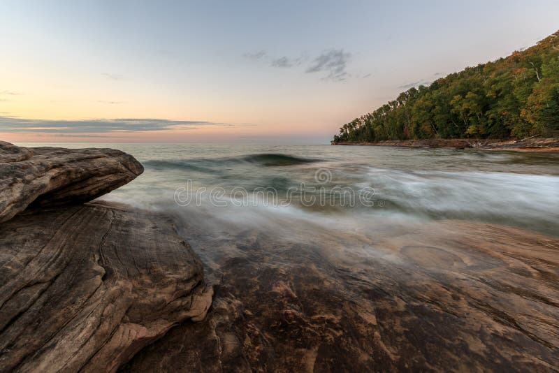 Sunset hues color the sky and autumn colors dot the forest at Miners Beach in the Upper Peninsula of Michigan. Easy waves ebb across the sandstone rock formations that have been here for centuries. Sunset hues color the sky and autumn colors dot the forest at Miners Beach in the Upper Peninsula of Michigan. Easy waves ebb across the sandstone rock formations that have been here for centuries.