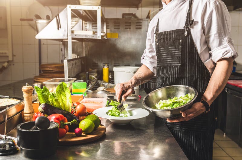 Professional chef cooking in the kitchen restaurant at the hotel, preparing dinner. A cook in an apron makes a salad of vegetables and pizza. Professional chef cooking in the kitchen restaurant at the hotel, preparing dinner. A cook in an apron makes a salad of vegetables and pizza