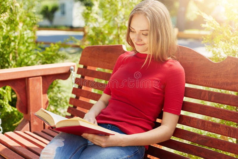 Beruhigende Frau mit dem angemessenen Haar, das rote Strickjacke und die Jeans sitzen an der hölzernen großen Bank hält Buchlesun stockfotografie Beruhigende Frau mit dem angemessenen Haar, das rote Strickjacke und die Jeans sitzen an der hölzernen großen Bank hält Buchlesun stockfotografie