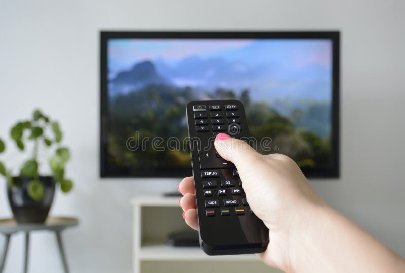 ?berwachender Fernsehapparat . Eine Hand, die die Fernbedienung mit einem Fernseher im Hintergrund hält stockfoto