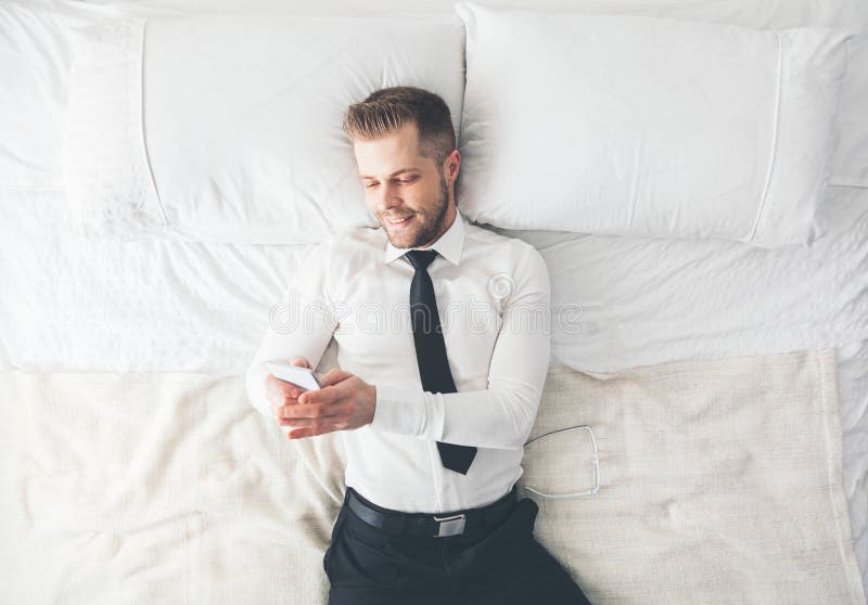 Beschneidungspfad eingeschlossen Hübscher Geschäftsmann, der auf dem Bett simst von seinem Smartphone liegt lizenzfreie stockfotografie Beschneidungspfad eingeschlossen Hübscher Geschäftsmann, der auf dem Bett simst von seinem Smartphone liegt lizenzfreie stockfotografie