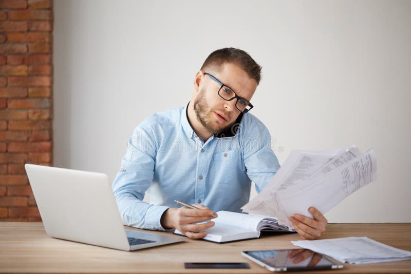 Beschäftigter starker unshaved Geschäftsmann in den Gläsern und in Hemd, die in einem bequemen hellen Büro, durch schauend sitzt stockbild Beschäftigter starker unshaved Geschäftsmann in den Gläsern und in Hemd, die in einem bequemen hellen Büro, durch schauend sitzt stockbild