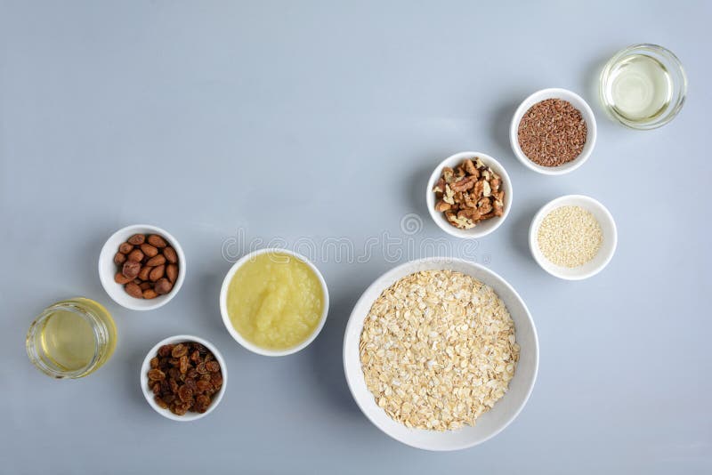 Bestandteile für das Kochen des selbst gemachten Granolas: Hafermehl-, Apfelmus-, Honig-, Öl-, Mandel-, Walnuss-, Rosinen-, Flachs- und Sesamsamen auf hölzernem grauem Hintergrund Flache Lage Beschneidungspfad eingeschlossen