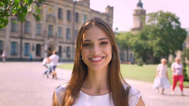 Bevindt de jonge vrouw van Nice zich in park in dag, lettend op bij camera, het glimlachen, mensengang op achtergrond