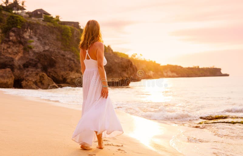 Beztroska kobieta w biel sukni odprowadzeniu na plaży przy zmierzchem fotografia stock Beztroska kobieta w biel sukni odprowadzeniu na plaży przy zmierzchem fotografia stock