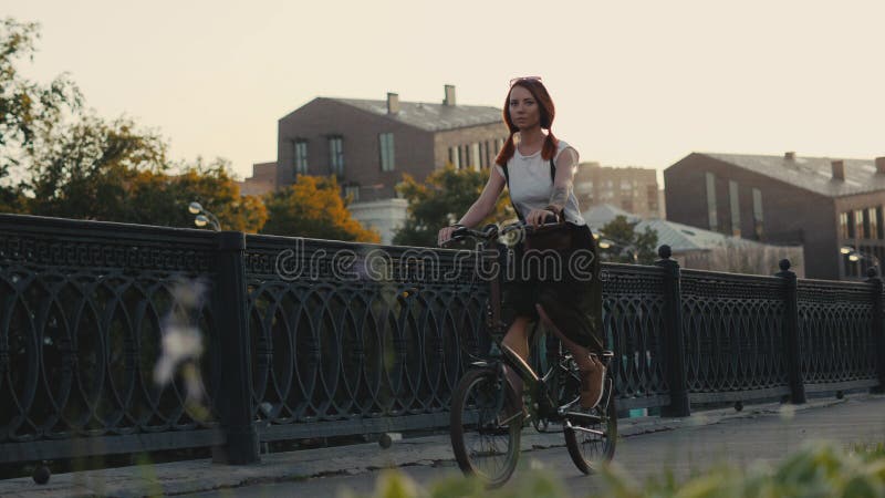 Bicicleta de cabelo vermelha da equitação da mulher na cidade na construção da fachada do fundo fotografia de stock Bicicleta de cabelo vermelha da equitação da mulher na cidade na construção da fachada do fundo fotografia de stock