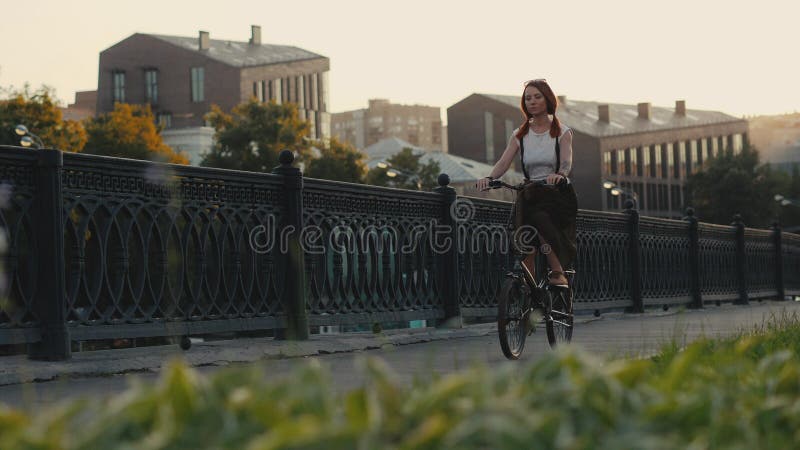 Bicicleta de cabelo vermelha da equitação da mulher na cidade na construção da fachada do fundo fotografia de stock Bicicleta de cabelo vermelha da equitação da mulher na cidade na construção da fachada do fundo fotografia de stock