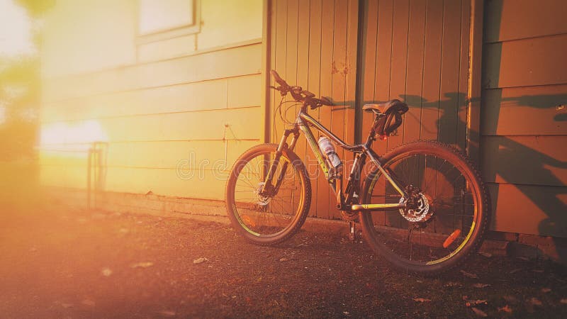 A rugged mountain bike leans against the doorway of an old shed under the pretty sunset lighting during a summer evening. A rugged mountain bike leans against the doorway of an old shed under the pretty sunset lighting during a summer evening.