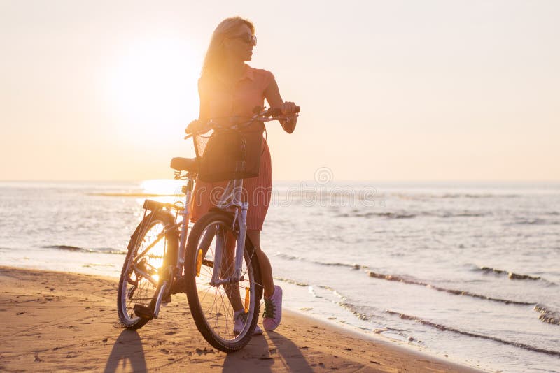 Bicicletta di guida della donna alla moda sulla spiaggia al tramonto