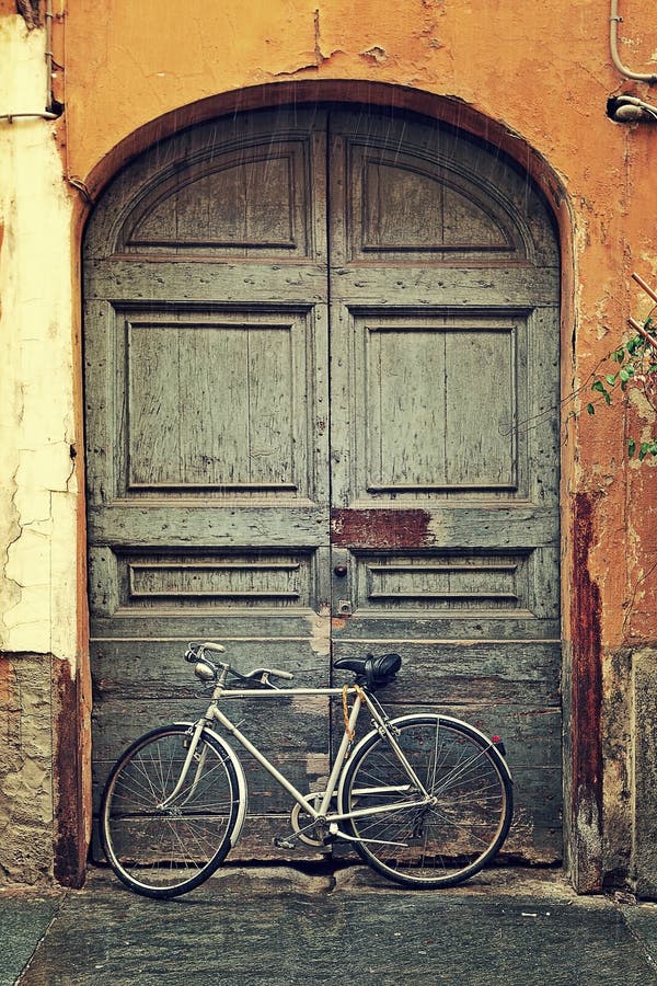 Vertical oriented image of bicycle leaning against old wooden door at the entrance to house on rainy day in Alba, Italy. Vertical oriented image of bicycle leaning against old wooden door at the entrance to house on rainy day in Alba, Italy.