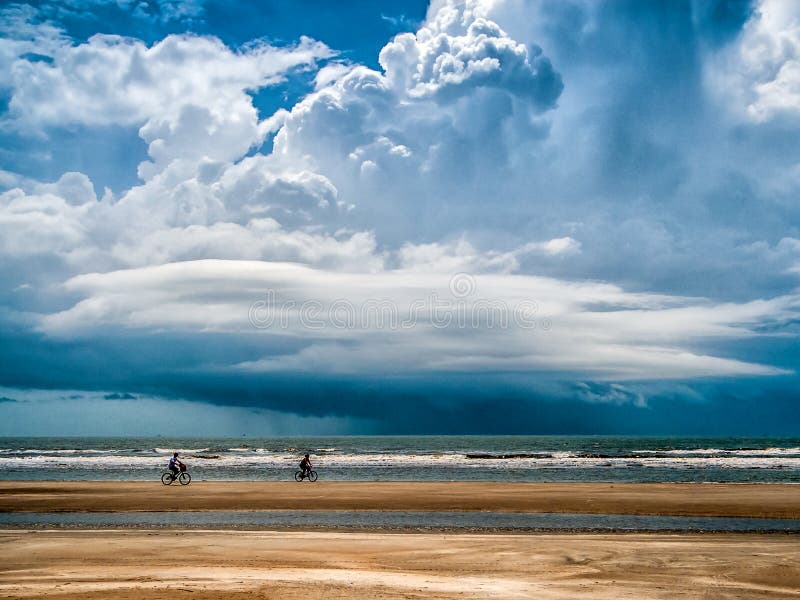 Two vacationers enjoy bicycling on a beach shortly before a large storm arrives from the sea. Two vacationers enjoy bicycling on a beach shortly before a large storm arrives from the sea.