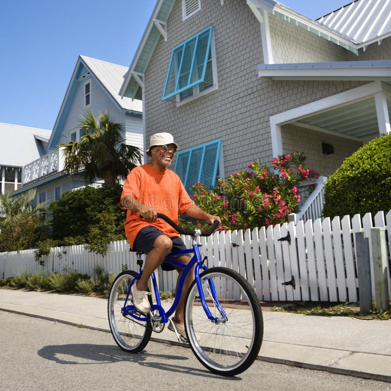 Middle aged African American man bicycling down street next to homes and fence. Middle aged African American man bicycling down street next to homes and fence.