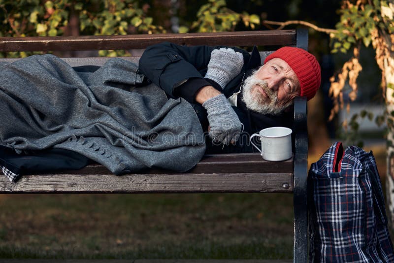 Roofless male lying on street bench asking for money, for any help. Lagguage near bench. Desperate and lonely homeless man without shelter. Roofless male lying on street bench asking for money, for any help. Lagguage near bench. Desperate and lonely homeless man without shelter