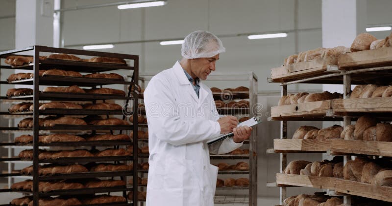 In a big bakery industry main worker checking the shelves full of fresh baked bread make notes on his map in a big stock photos