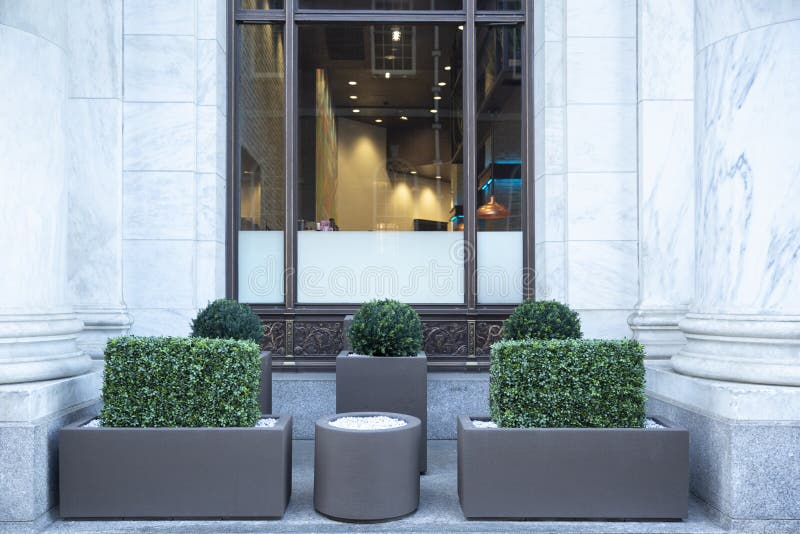Big gray, metal planters with foliage plants and terrace design with a modern mix of construction material. Bushes against a. Building with big window